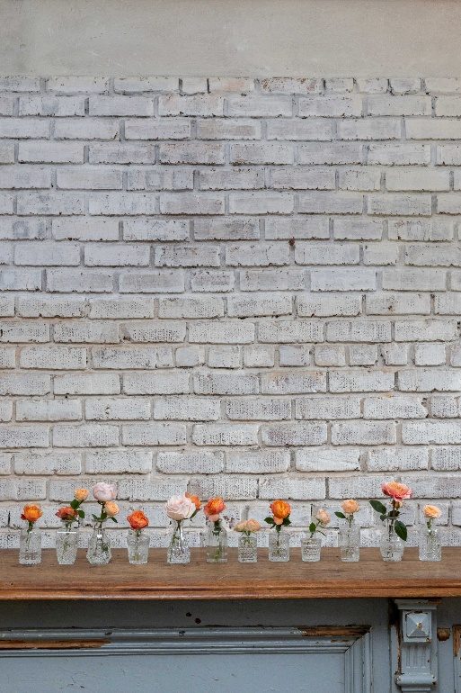 Twelve clear bud vases in a row on a wooden mantel, each holding a single peach or pink rose
